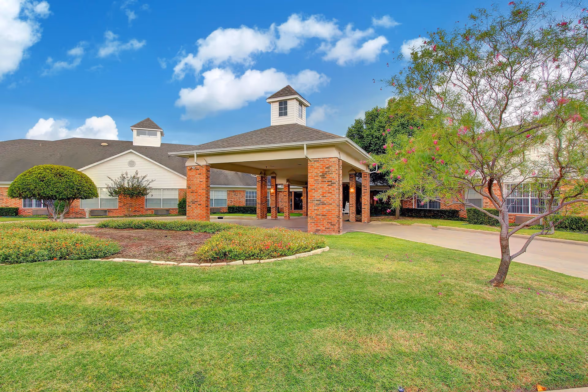 Exterior view of Crestwood Health and Rehabilitation Center showing a covered entrance with brick pillars, a well-maintained lawn, landscaped bushes, and a tree with pink flowers under a blue sky with scattered clouds.
