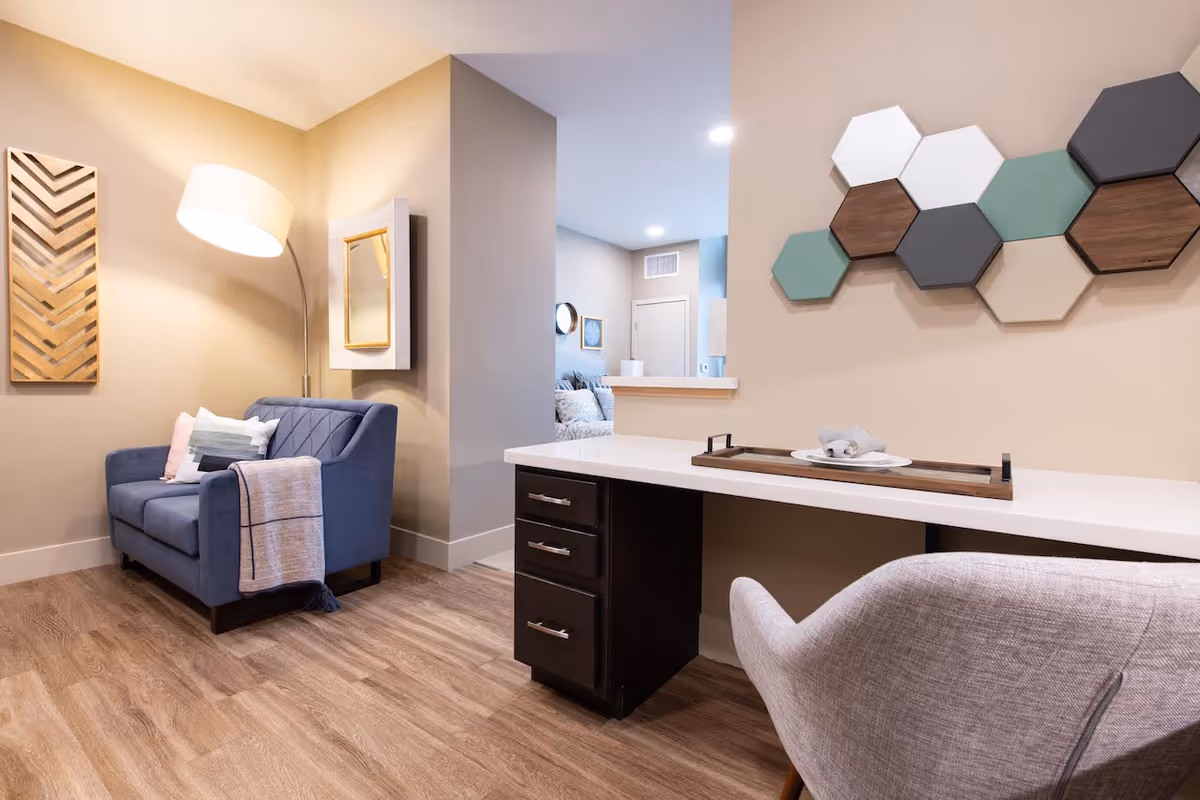 A cozy interior space featuring a blue upholstered loveseat with pillows and a throw blanket, a tall floor lamp, a wall-mounted mirror, and a decorative wooden wall art. There is a white desk with dark drawers and a gray chair, with a tray holding a plate and napkins on the desk. The room has wood flooring and beige walls, with a glimpse into an adjacent room showing a bed and wall decor.