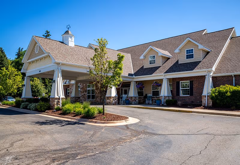 Exterior view of a senior living facility building with a covered entrance, brick and stone facade, dormer windows, and landscaping including a small tree and bushes under a clear blue sky.