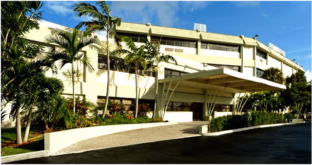 Exterior front view of a multi-story building with a covered entrance, surrounded by palm trees and landscaping under a blue sky.