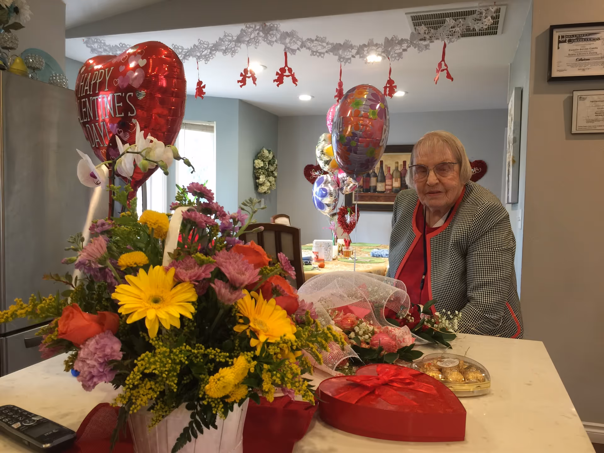 An elderly woman sitting at a table decorated with Valentine's Day themed items including a heart-shaped balloon, colorful flowers, a heart-shaped box of chocolates, and other balloons in a cozy indoor setting.