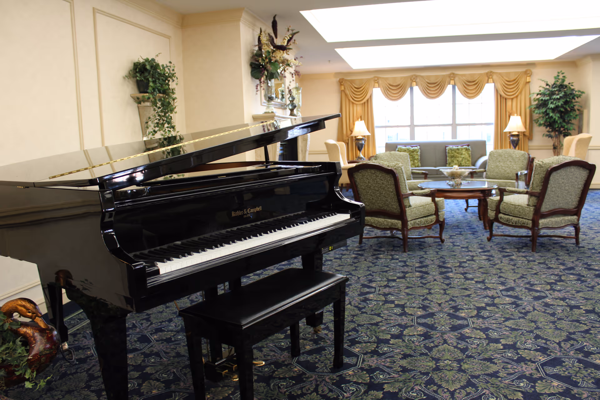 A spacious senior living community room featuring a black grand piano with a matching bench in the foreground. The room has patterned blue and green carpet, several green upholstered armchairs arranged around a wooden coffee table, a sofa with green cushions, two table lamps, large windows with gold curtains, and decorative plants.