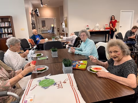Several elderly residents sit around a communal dining table eating slices of watermelon in a senior living activity room.