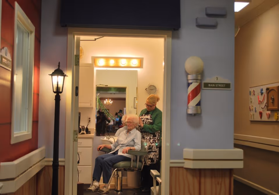 An elderly woman is seated in a salon chair inside a hair salon area, getting her hair done by a stylist. The salon is decorated with a barber pole on the wall and a sign that reads 'Main Street'. The scene is viewed through an open doorway from a hallway with warm lighting and decorative elements.