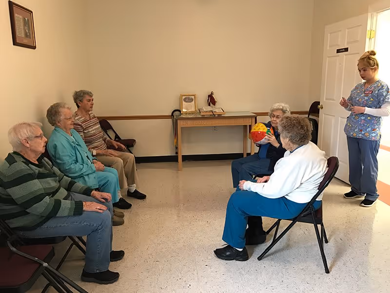 A group of elderly women sitting in chairs arranged in a semi-circle in a plain room with beige walls and a tiled floor. One woman is holding a colorful ball, and a caregiver in scrubs stands nearby observing. A small table with a few items is against the wall in the background.