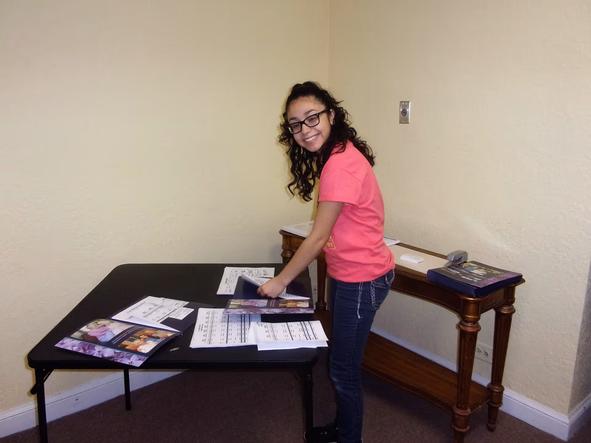 A young girl with curly hair and glasses, wearing a pink shirt and jeans, is standing and smiling while organizing papers on a black folding table in a room with beige walls and carpeted floor. There is a wooden console table against the wall with more papers and a stapler on it.