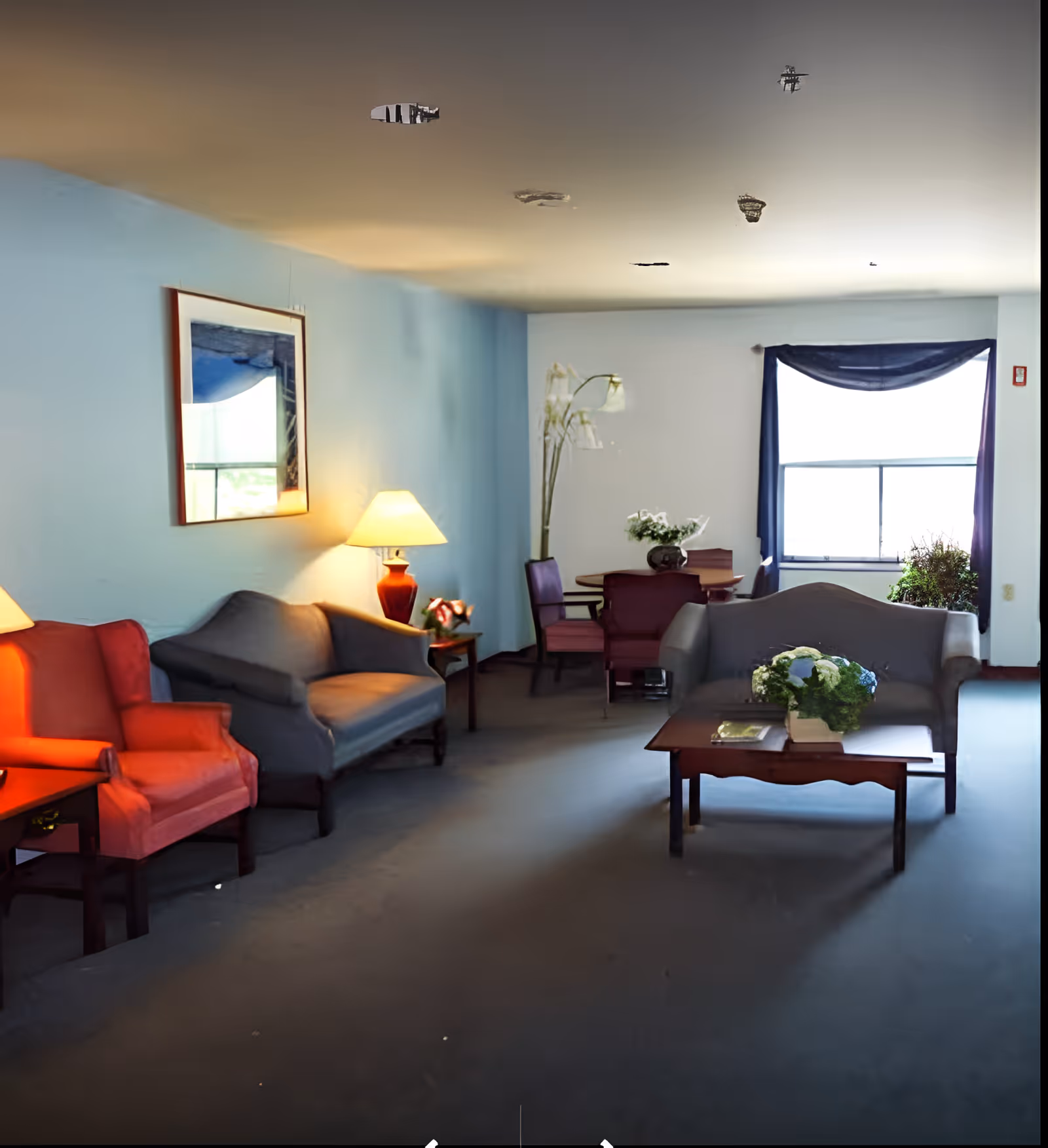A cozy living room area with a blue wall and a window with dark curtains. The room features a red armchair, two gray sofas, a wooden coffee table with a flower arrangement, a side table with a lamp, and a round dining table with chairs and a tall flower vase. The room is softly lit by natural light from the window and the lamp.