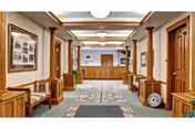 Spacious interior hallway of a retirement center with wooden columns and trim, carpeted floor with patterned rugs, chairs along the walls, framed pictures on the left wall, and a reception desk at the far end under ceiling lights.
