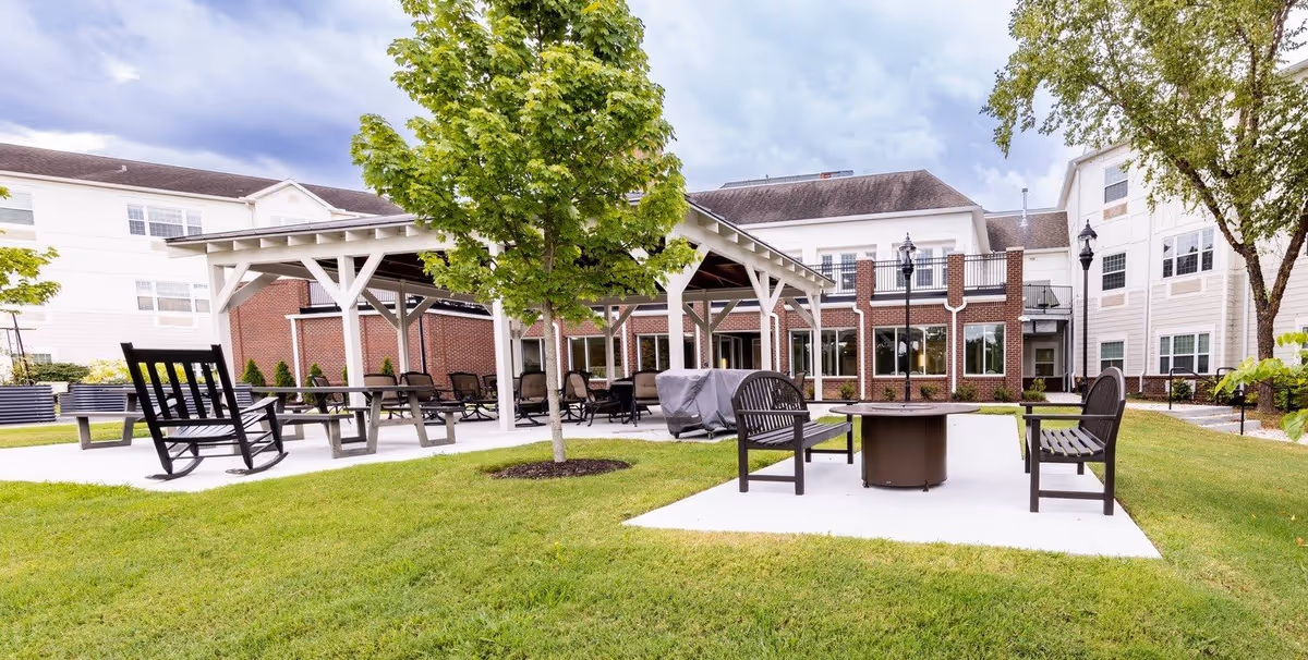 Outdoor seating area at Mary Queen of Angels Assisted living facility featuring a covered patio with multiple chairs and tables, a fire pit surrounded by benches, green grass, and trees with the building in the background under a cloudy sky.
