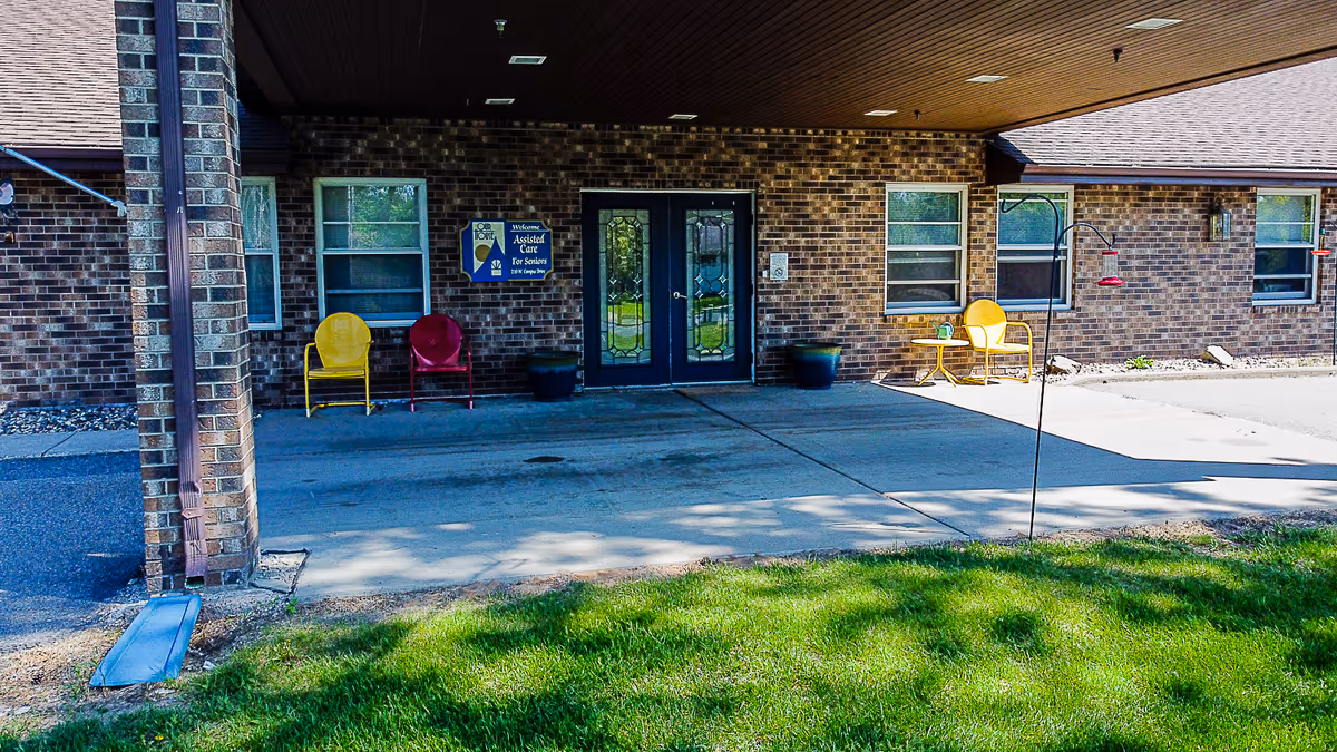 Covered brick entrance to a senior living facility with double decorative doors, colorful chairs, and a small lawn in front.
