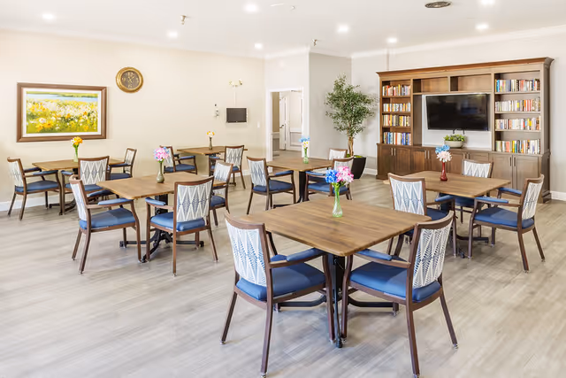 A bright and spacious common area with multiple wooden tables and chairs arranged neatly. Each table has a small vase with colorful flowers. There is a large wooden bookshelf with a flat-screen TV mounted in the center. The room has light-colored walls, a clock, a framed painting, and a potted plant near the bookshelf. The floor is covered with light wood-style flooring.