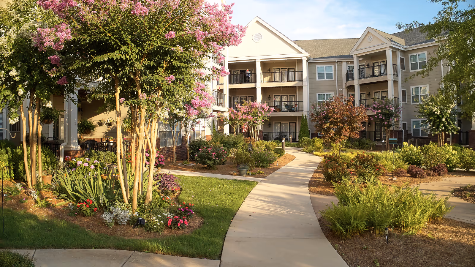 A landscaped outdoor courtyard area at Fairview At Redstone Village featuring a winding concrete pathway surrounded by green grass, flowering bushes, and trees with pink and white blossoms. In the background, there is a three-story residential building with balconies and large windows under a partly cloudy sky.