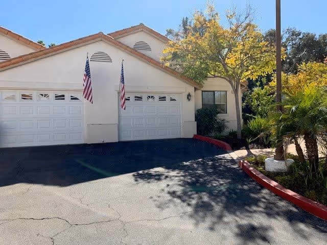 Exterior view of a residential building with two white garage doors, two American flags mounted on the wall between the garages, a tree with yellow leaves, and surrounding greenery including palm plants. The driveway is paved and bordered by a red curb.