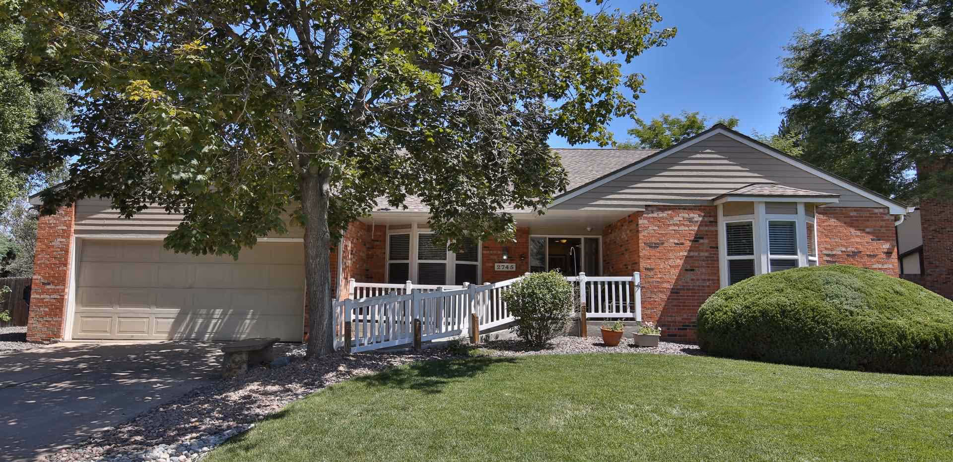 Front exterior view of a single-story brick house with a two-car garage, a white railing ramp leading to the front door, a large tree providing shade, and well-maintained green lawn and bushes under a clear blue sky.