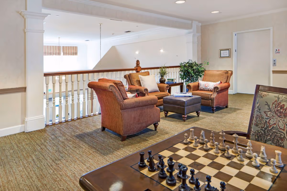 Upholstered armchairs and an ottoman arranged around a table in a bright communal lounge area with a chessboard in the foreground.