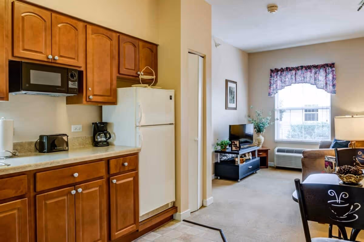 Interior view of a senior living facility apartment showing a kitchen area with wooden cabinets, a microwave, toaster, coffee maker, and refrigerator. Adjacent to the kitchen is a living room with a window covered by a floral valance, a TV on a stand, a sofa, and a small dining table with chairs.