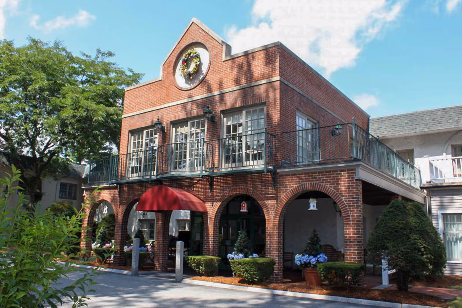 Exterior view of a two-story brick building with arched openings on the ground floor and a balcony with railing on the upper floor. The building has a red awning over the entrance and a decorative wreath on the upper facade. There are shrubs, potted plants, and trees around the building under a partly cloudy blue sky.