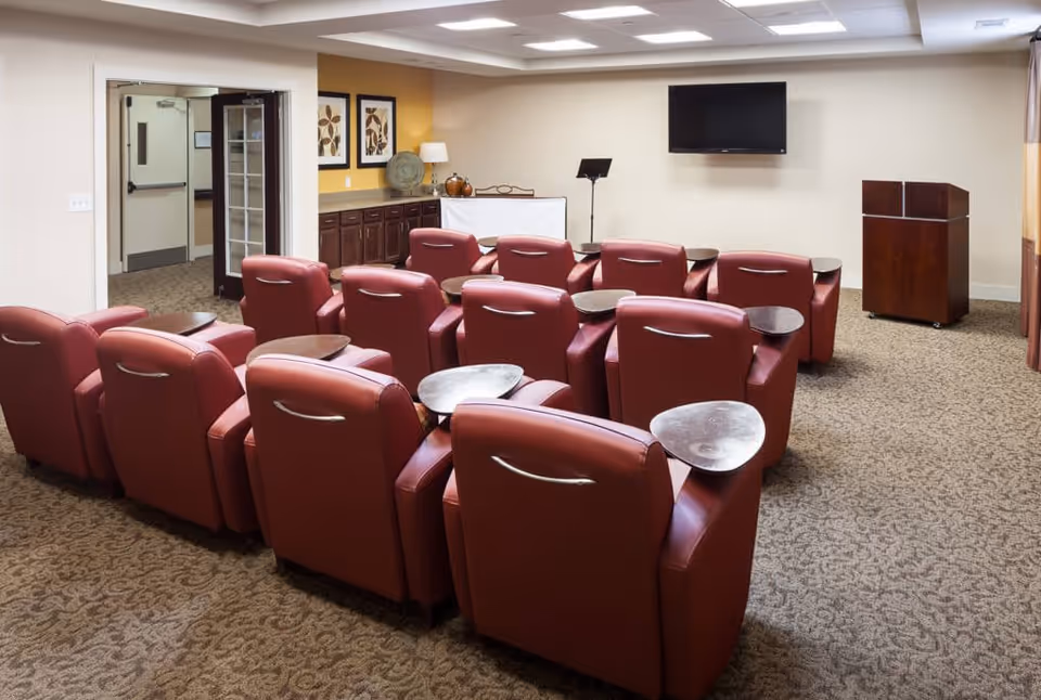 A small theater or presentation room with two rows of red leather recliner chairs, each equipped with a small attached table. At the front of the room is a podium and a flat-screen TV mounted on the wall. The room has beige walls, carpeted floor, and a side cabinet with decorative items and framed artwork above it.