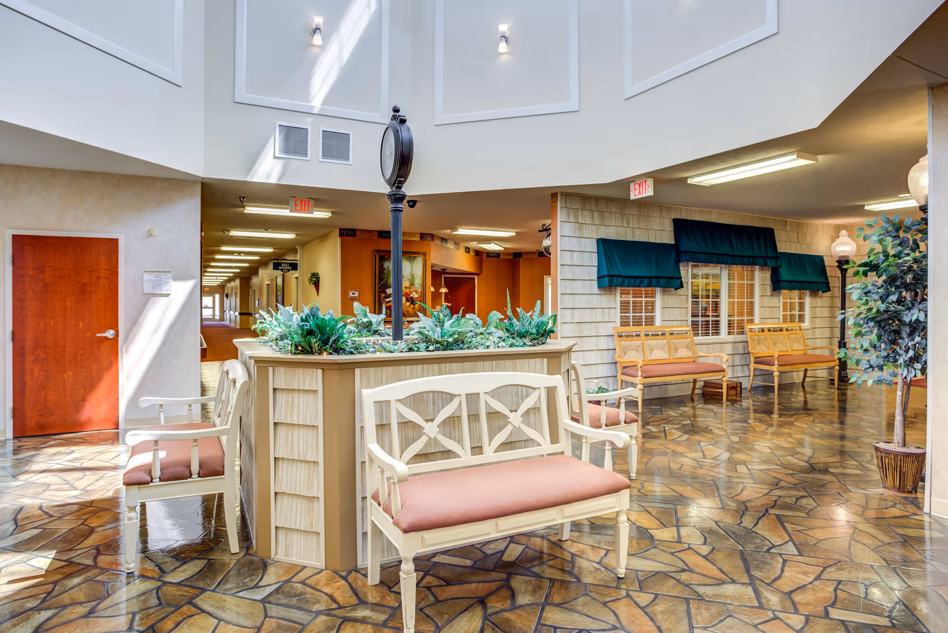 A bright and spacious indoor common area in a senior living facility featuring a central planter with green plants and a tall clock. Surrounding the planter are white wooden benches with pink cushions. The floor has a patterned tile design, and there are additional wooden benches with orange cushions along the walls. The walls have windows with green awnings, and there are potted plants and lamps adding to the decor. The ceiling is high with skylights allowing natural light to enter.