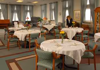Bright dining room with round tables, white tablecloths, flowers, and staff in background.