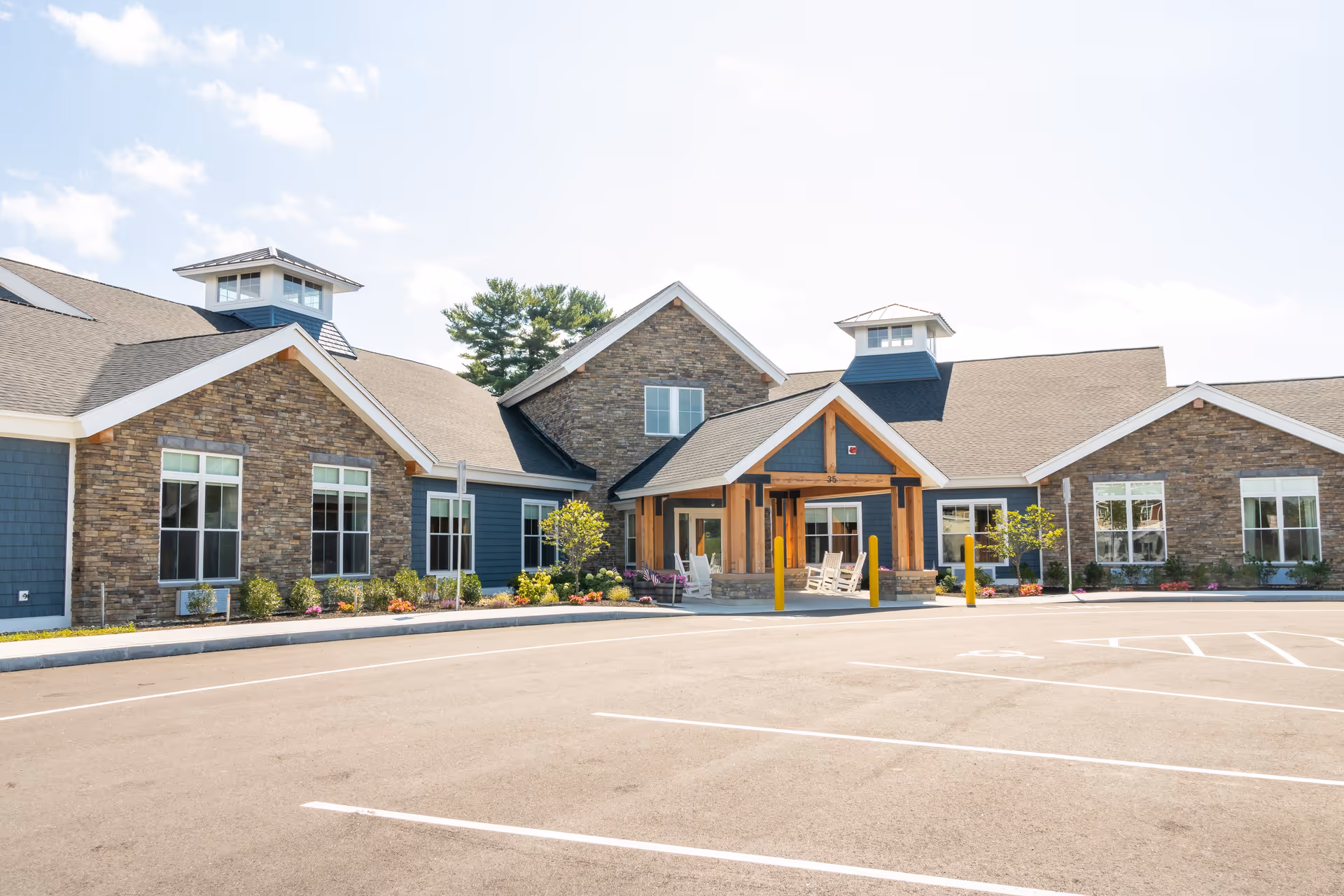 Exterior view of a single-story senior living facility building with stone and blue siding, multiple windows, a covered entrance with wooden beams, rocking chairs on the porch, and a spacious parking lot in front under a partly cloudy sky.