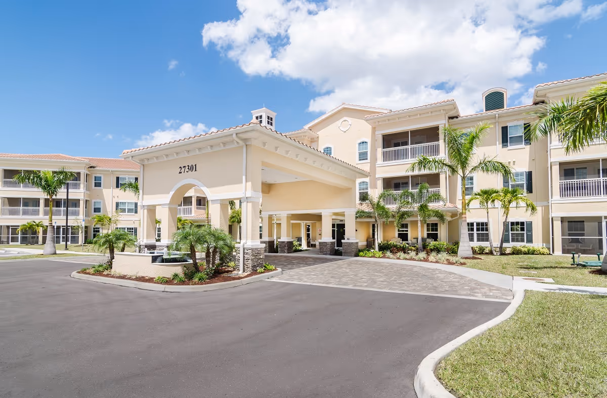 Exterior view of American House Bonita Springs senior living facility showing a large beige building with multiple balconies, palm trees, a covered entrance with the number 27301, and a clear blue sky with some clouds.