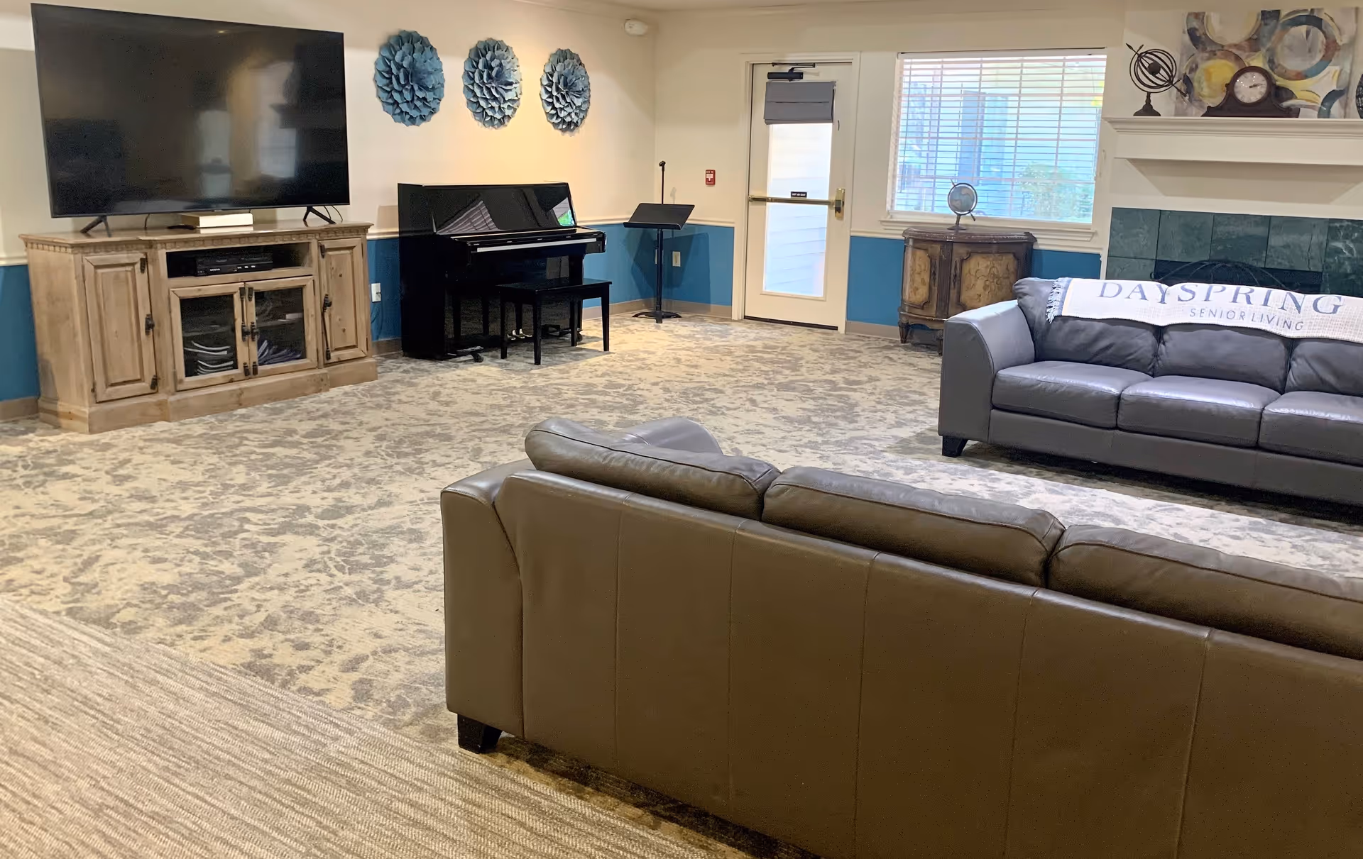 A senior living facility common area with two leather sofas, a large flat-screen TV on a wooden cabinet, a black upright piano, and decorative blue wall art. There is a window with blinds, a door, and a fireplace with decorative items on the mantel. A blanket with the text 'DaySpring Senior Living' is draped over one sofa.