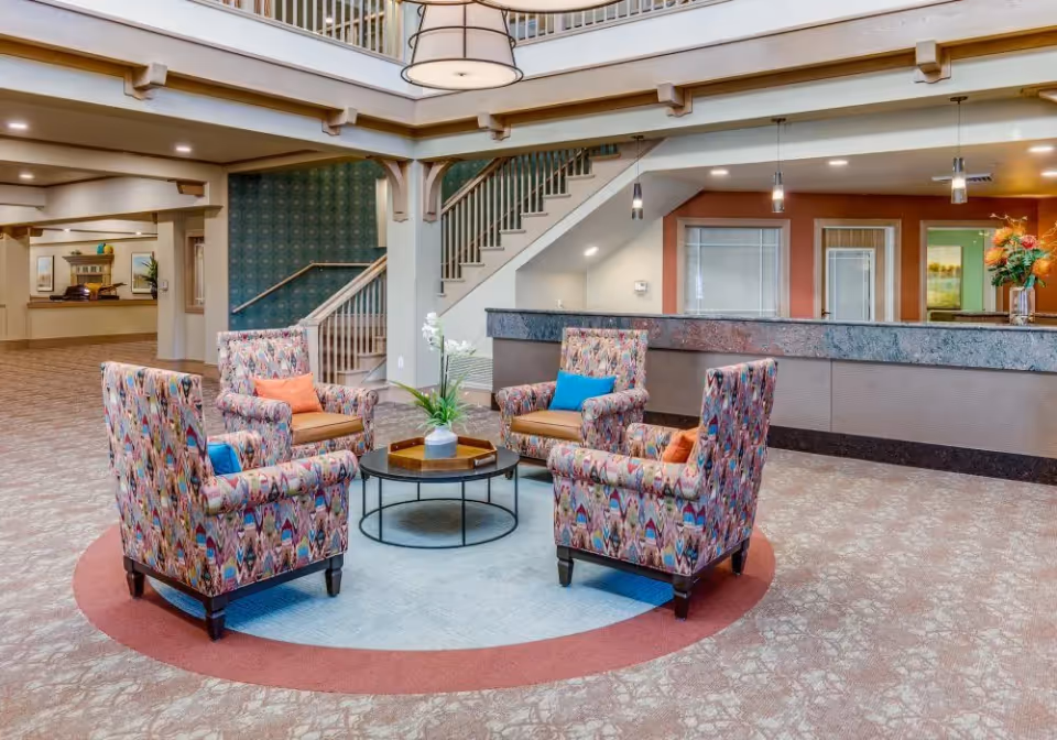 A spacious and well-lit lobby area with four colorful patterned armchairs arranged around a round coffee table with a plant centerpiece. Behind the seating area is a long reception desk with pendant lights hanging above. A staircase with a wooden handrail leads to an upper floor, and the walls are decorated with a mix of neutral and accent colors.