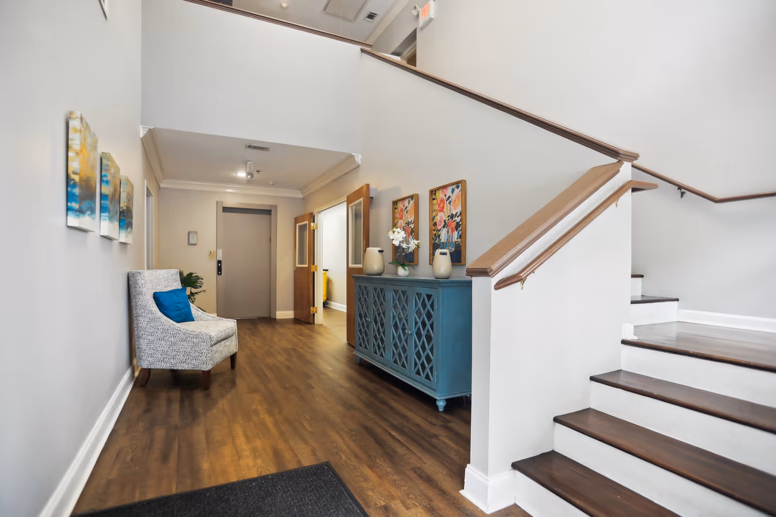 Well-lit interior entry hall with a staircase, teal console table, artwork, and a patterned chair.