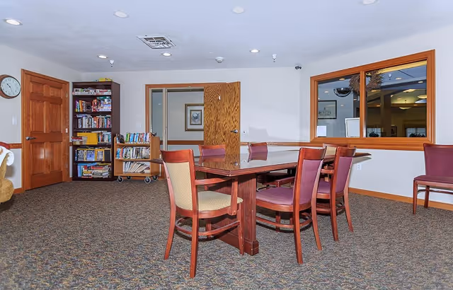 A communal room with a rectangular wooden table surrounded by chairs, a bookshelf with games and books, and a window into an adjacent area.