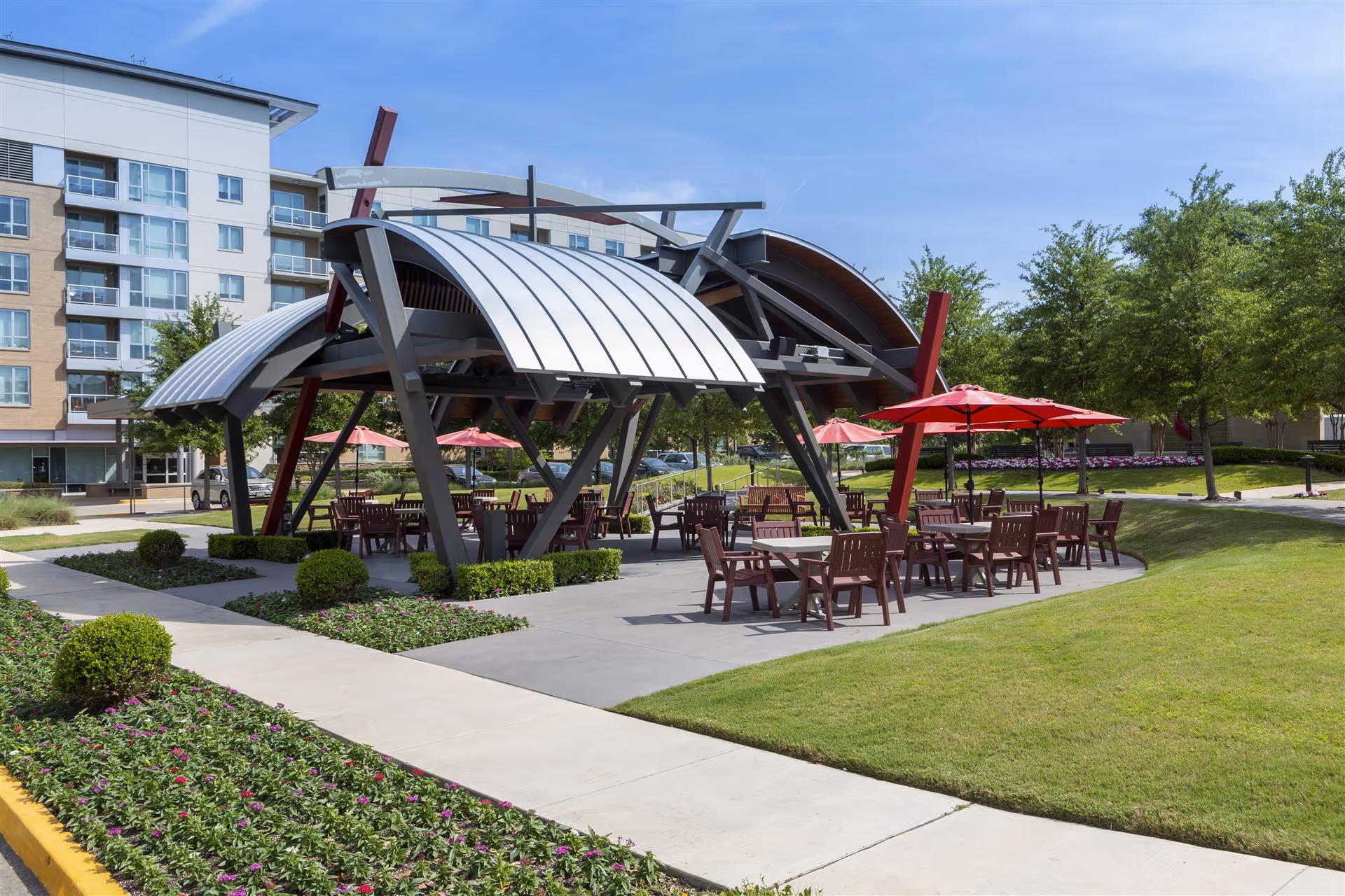 Outdoor seating area with modern metal and wood pavilion structure, multiple tables and chairs with red umbrellas, surrounded by green grass, trees, and flower beds, with a multi-story building in the background under a clear blue sky.