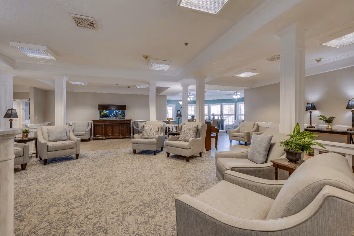 A spacious and well-lit living room area in a senior living facility featuring multiple beige upholstered armchairs arranged in a circular pattern on a patterned carpet. The room has white columns, a wooden cabinet with a fish tank on top, several side tables with lamps and plants, and large windows with white shutters allowing natural light to fill the space.