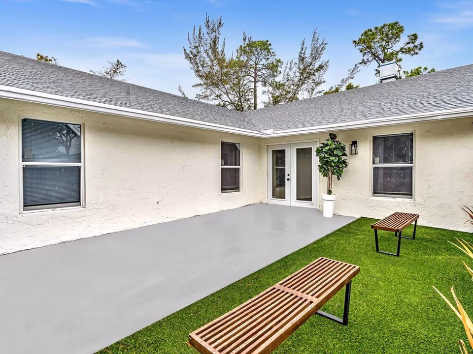 Outdoor courtyard area with two wooden benches on artificial grass, a concrete patio, a potted plant near double glass doors, and windows on the beige exterior walls of a single-story building under a blue sky.