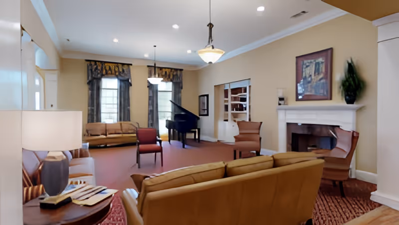 A spacious living room with beige walls and carpeted floor featuring multiple seating options including sofas and armchairs. There is a grand piano near two tall windows with dark curtains, a white fireplace with a framed painting above it, and a small table with a lamp and books in the foreground.