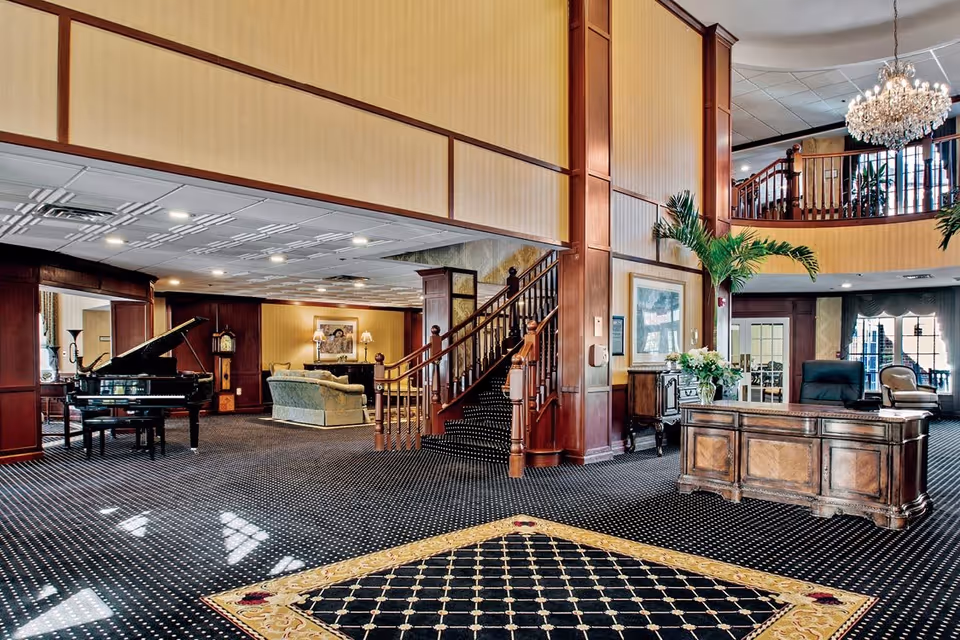 Spacious and elegant interior of a senior living facility featuring a grand piano, a wooden staircase with carpeted steps, a chandelier, a wooden reception desk with a chair, potted plants, and comfortable seating area with sofas and lamps in the background.