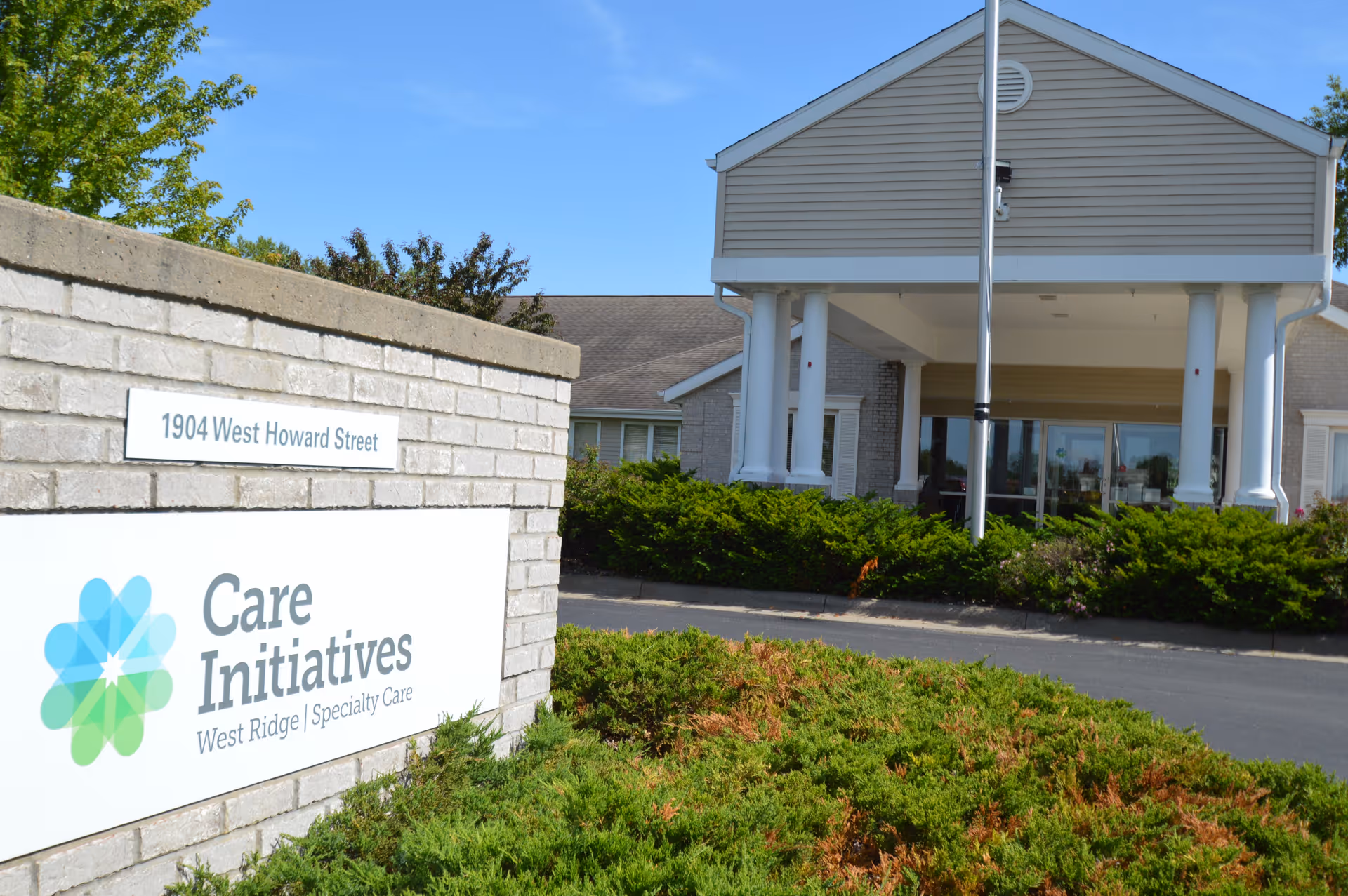 Exterior view of the entrance to Care Initiatives West Ridge Specialty Care facility with a sign displaying the address 1904 West Howard Street and the facility name. The building has a covered entryway supported by white columns, surrounded by greenery and bushes under a clear blue sky.