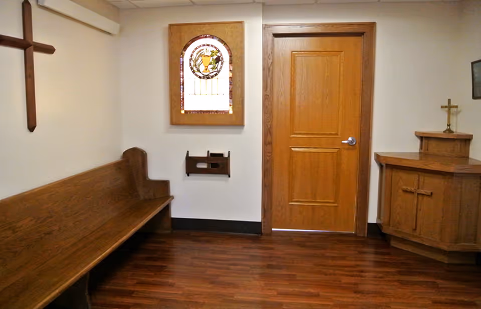 A small chapel room with wooden pew seating on the left, a wooden door in the center, and a wooden altar with a cross on the right. A stained glass window with a religious symbol is mounted on the wall above a wooden holder. The floor is dark wood, and the walls are light-colored.