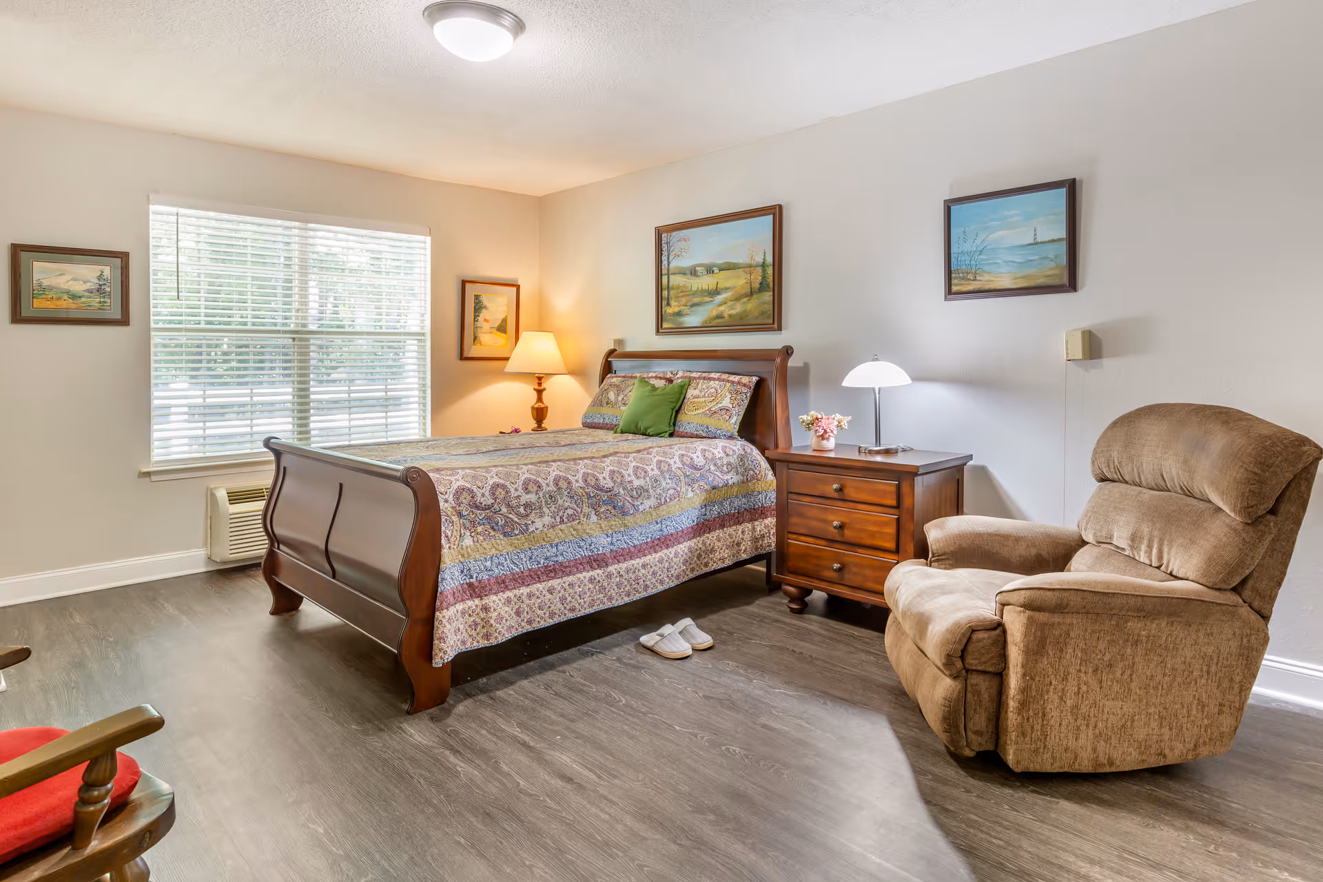 A cozy bedroom with a wooden bed featuring a colorful quilt and a green pillow. Next to the bed is a wooden nightstand with a lamp and a small flower vase. A comfortable brown recliner chair is positioned nearby. The room has a large window with blinds, three framed landscape paintings on the walls, and a wooden rocking chair with a red cushion partially visible in the corner.