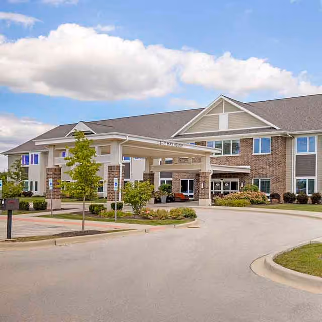 Entrance and covered porte-cochère of a two-story brick senior living facility with driveway and landscaped grounds under a partly cloudy sky.