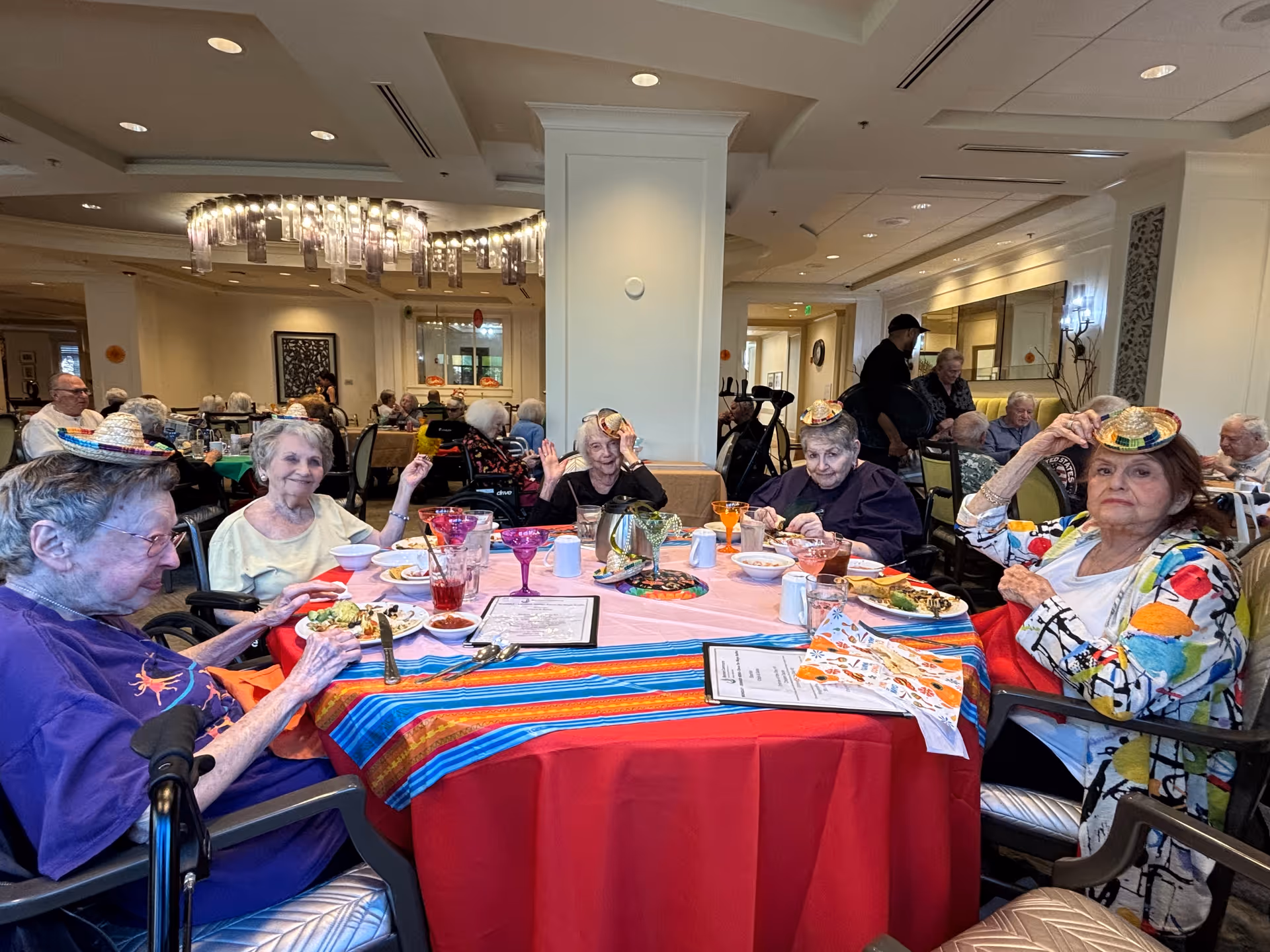A group of elderly residents seated around a decorated dining table in a communal dining room wearing small festive hats.