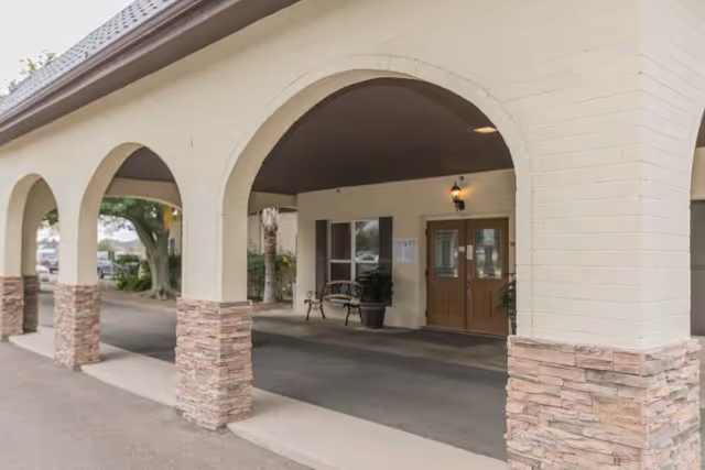 Covered entrance area of a building with beige brick arches and stone bases, a bench, potted plants, and double wooden doors under a light fixture.