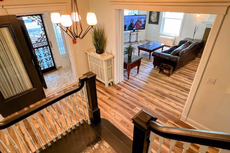 View from the top of a staircase looking down into a living room area with a brown leather couch, a coffee table, a side table with a plant, and a television mounted on the wall. The floor is wood with a natural finish, and there is a chandelier hanging above the staircase landing. The entrance door with decorative glass panels is visible to the left.