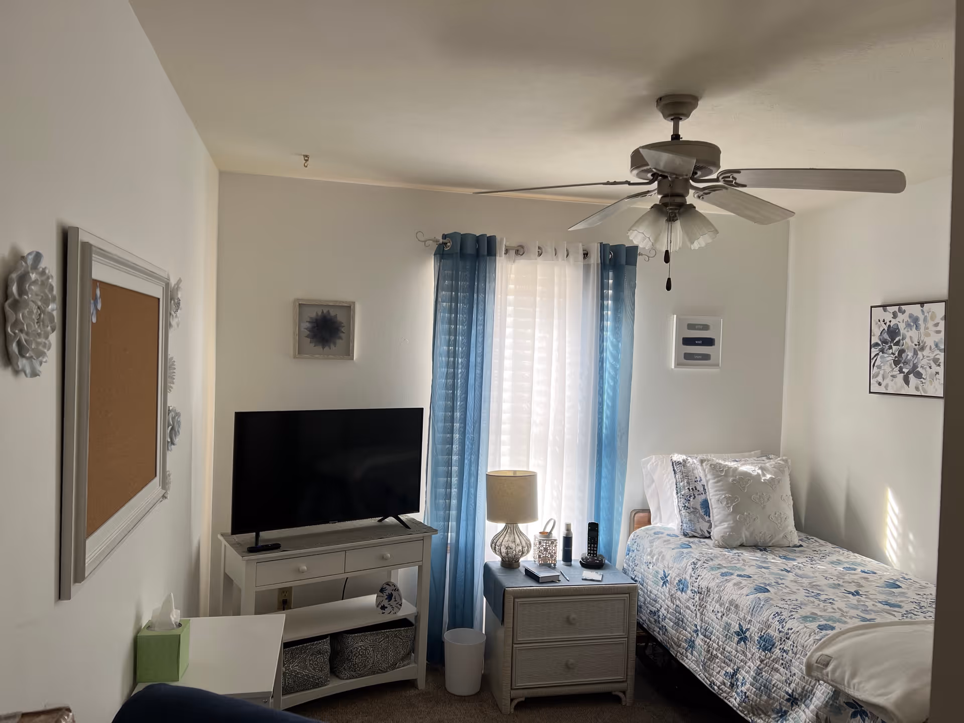 A small, cozy bedroom with a single bed covered in a blue and white floral quilt. There is a nightstand next to the bed with a lamp, a phone, and some small items. A flat-screen TV sits on a white stand against the wall. The window behind the nightstand has sheer white curtains with blue drapes. The room has light-colored walls with a few framed pictures and a ceiling fan with lights.