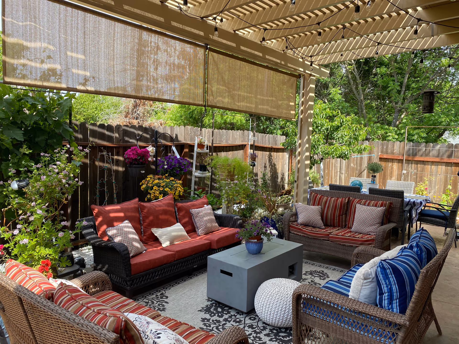 A cozy outdoor patio area with wicker furniture including sofas and chairs with colorful cushions in red, blue, and striped patterns. There is a rectangular coffee table in the center with a potted plant on it. The patio is covered with a pergola that has string lights hanging from it and roll-down shades partially lowered. The space is surrounded by a wooden fence and lush green plants and flowers.