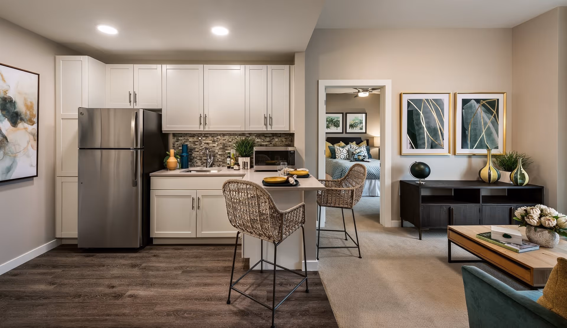 A modern kitchen and living area featuring a stainless steel refrigerator, stylish bar stools, and decorative elements. The space is well-lit and includes a view into a cozy bedroom with framed artwork on the walls.