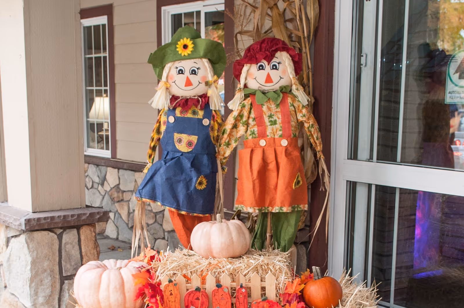 Two autumn scarecrow dolls standing among pumpkins and hay bales at the building's entrance.