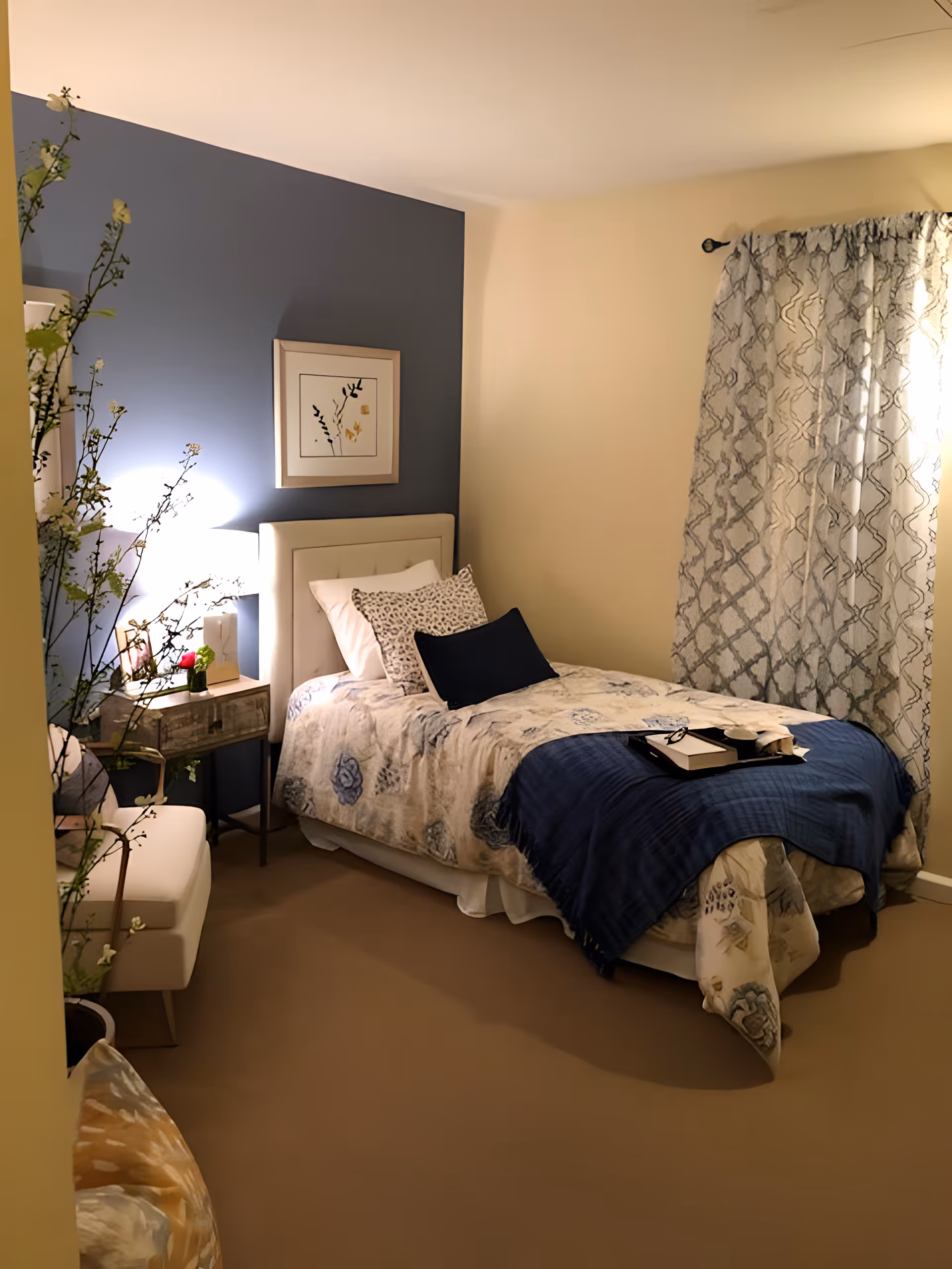 A cozy single bedroom with a bed against a blue accent wall, a nightstand with a lamp, patterned curtains, and decorative pillows.