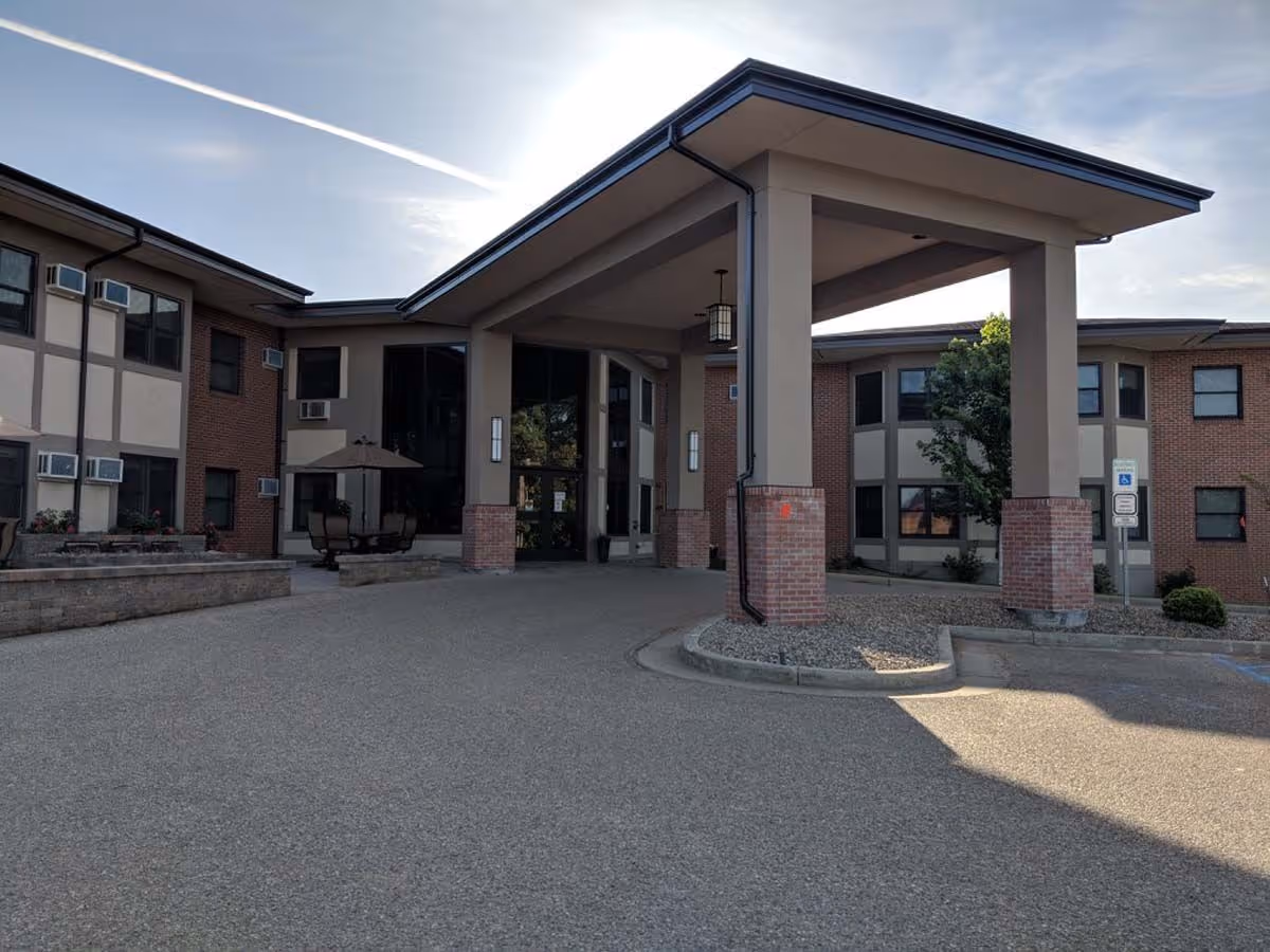 Exterior view of the Missouri Slope facility entrance featuring a covered drop-off area supported by large columns with brick bases, surrounded by a paved driveway and landscaped areas with shrubs and a tree.