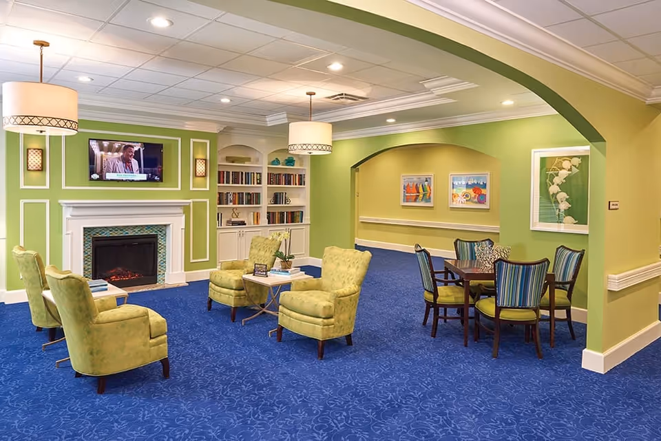 Bright communal living room with green armchairs and a fireplace, a TV and bookshelves, plus a dining table on blue carpet.
