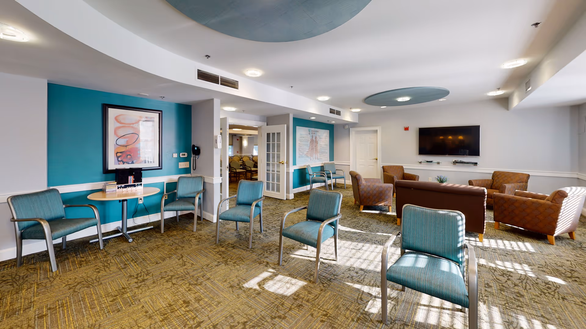 A spacious senior living facility common area with teal and brown chairs arranged in small groups. The room features a teal accent wall with framed artwork, a round table with a phone, a flat-screen TV mounted on a white wall, and large windows allowing natural light to fill the space. The ceiling has circular recessed lighting fixtures and a patterned carpet covers the floor.